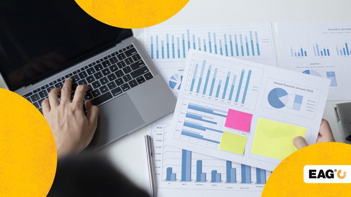 mãos de uma pessoa teclando no notebook e ao lado várias folhas de ofício com gráficos e dados da empresa