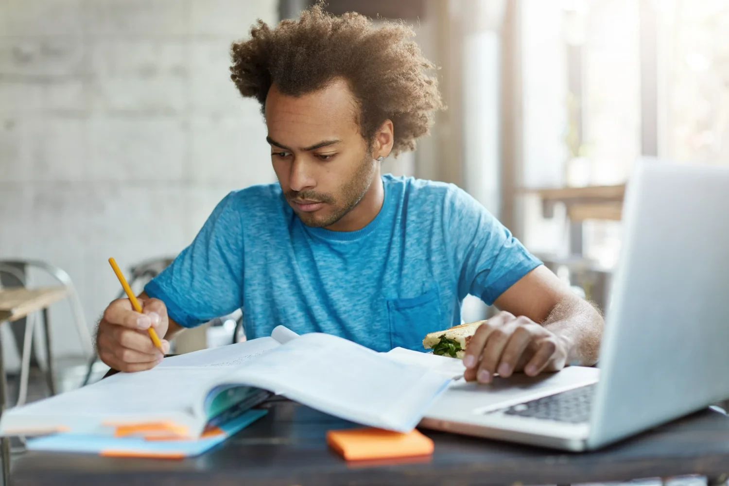Homem negro de camiseta azul estudando com livros e notebook em uma mesa representando a aprendizagem autodirigida.