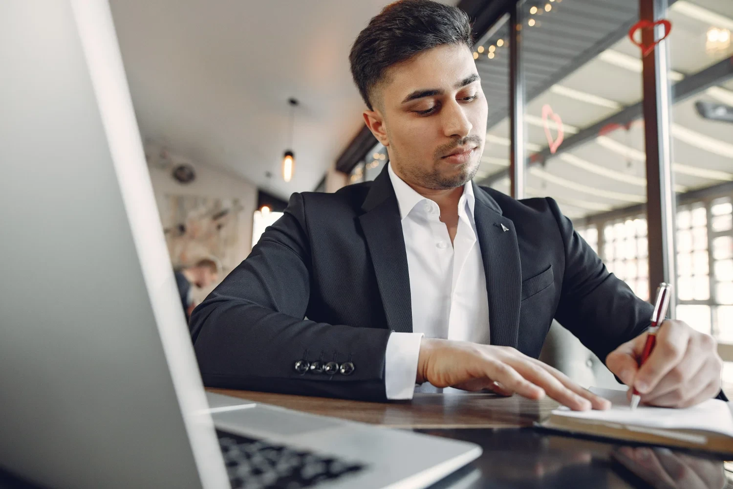 Homem branco de terno escrevendo em uma folha com notebook em frente representando um empresário estudando para sua capacitação gerencial.